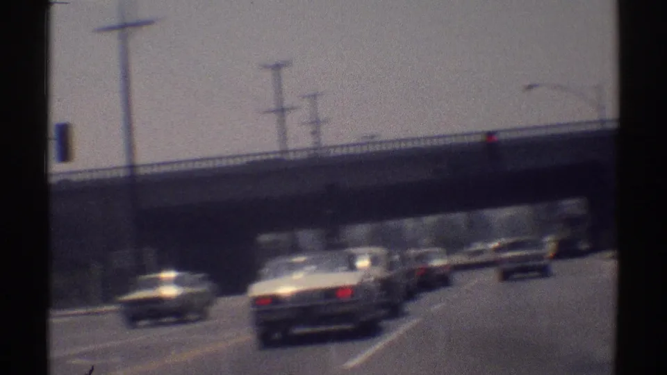 Moving-car view of 1969 Woodland Hills traffic passing under an overpass, with bright road glare and overhead poles.
