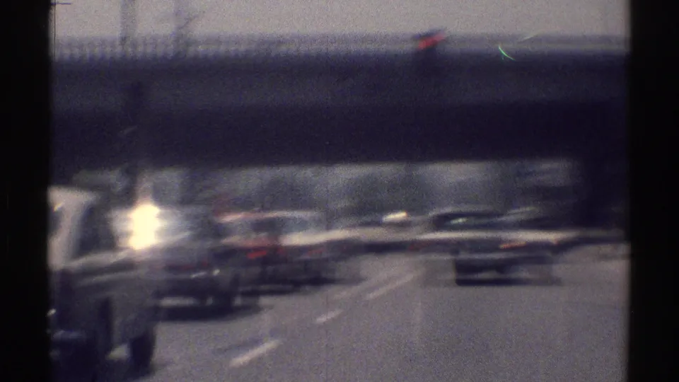 Late-1960s Woodland Hills driving view with cars under an overpass, sun flare, lane markings, and soft 8mm motion blur.