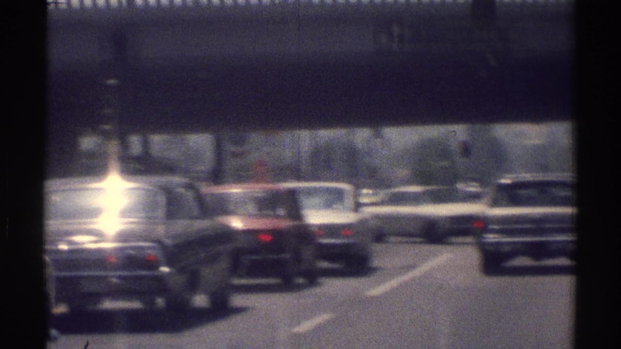 Traffic with 1960s cars moving under an overpass on a multi-lane highway.
