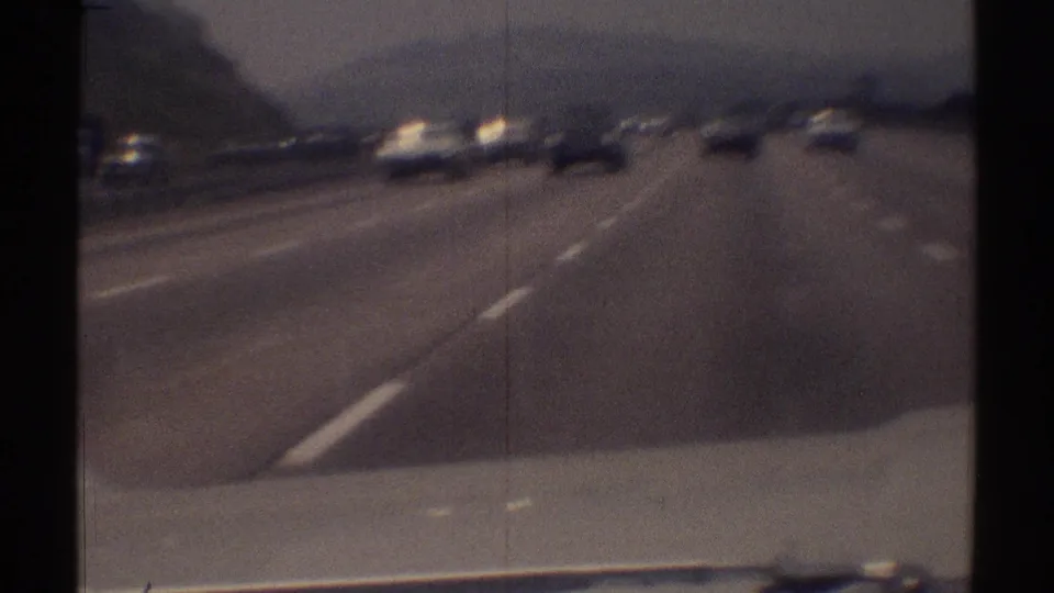 Moving-vehicle view of a Woodland Hills highway with 1960s traffic, lane markings, and a roadside hillside.