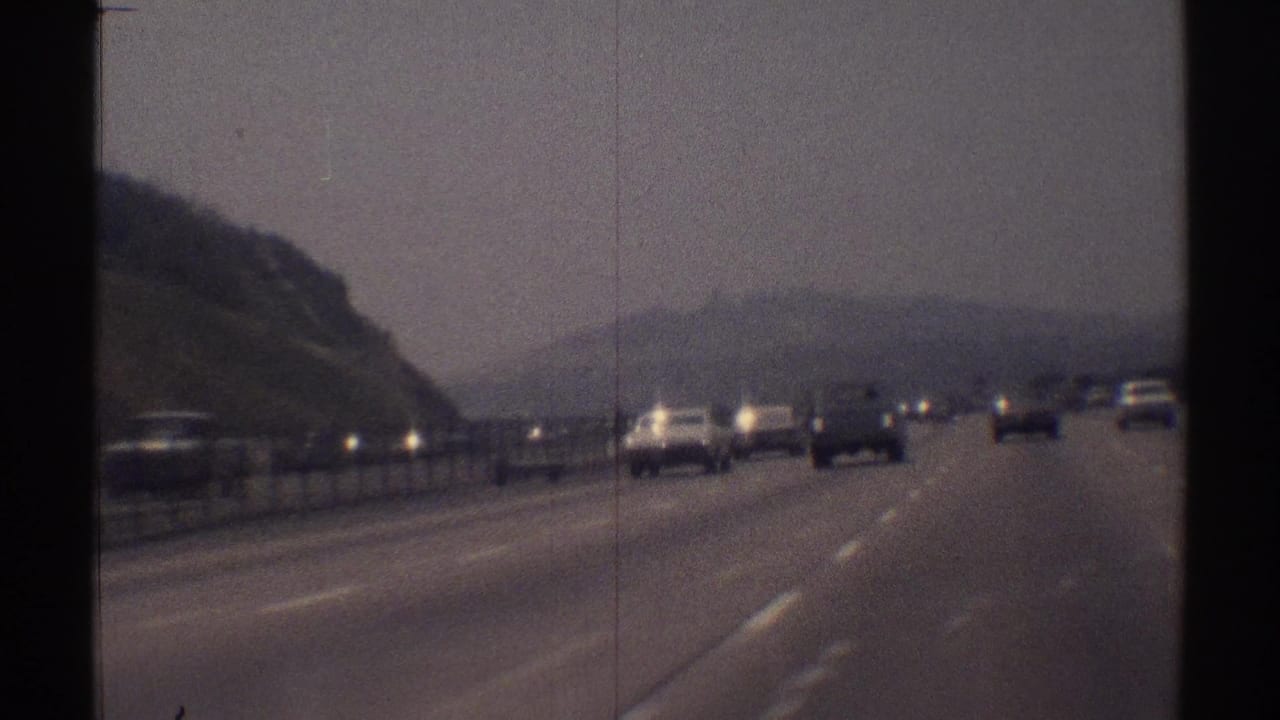 Cars travel on a multi-lane highway beside a hillside under a hazy sky.