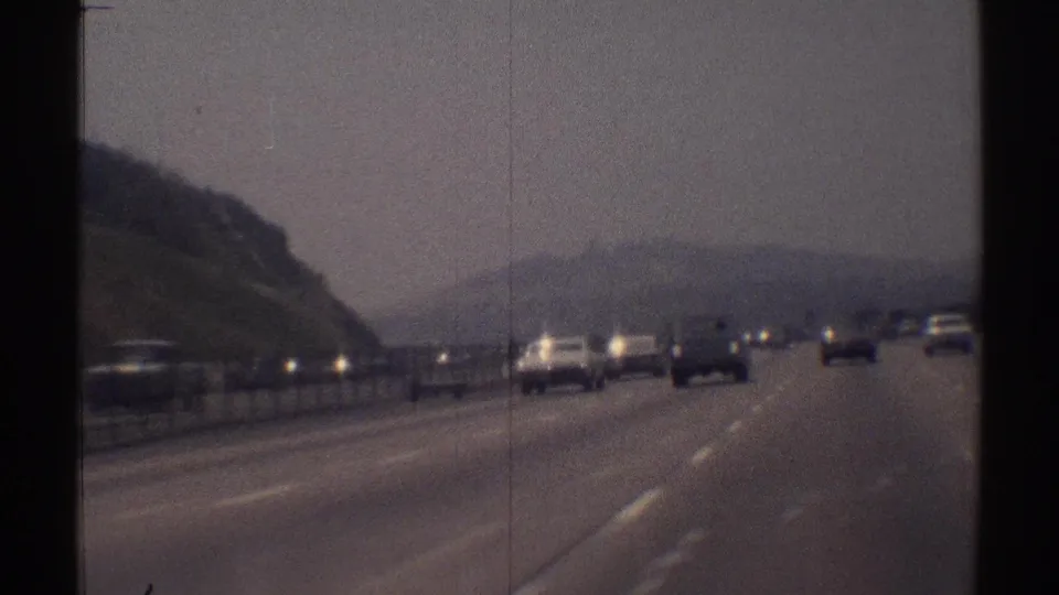 1969 Woodland Hills highway view from a moving vehicle, with cars ahead, a divider rail, hillside terrain, and a hazy sky.