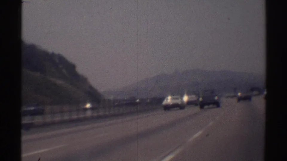 Moving 8mm view of cars on a divided Woodland Hills highway in 1969, with sloped hillsides and muted haze beyond the road.
