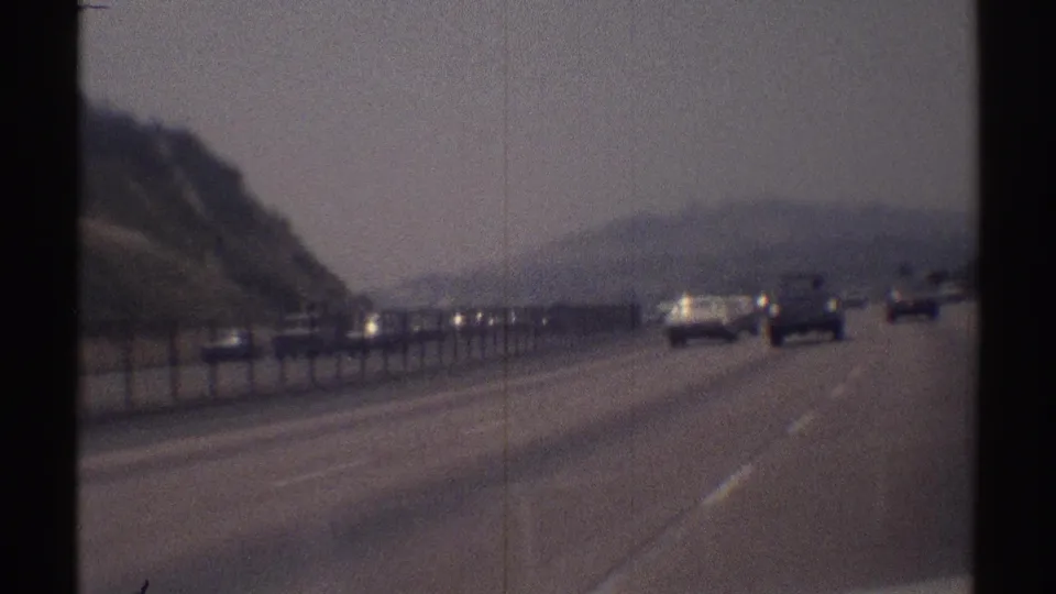 1969 8mm roadway view near Woodland Hills, showing cars ahead on a broad highway, a center barrier, hillside brush, and soft haze.