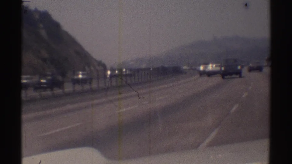 End frame of the 1969 Woodland Hills highway clip, with cars ahead, a visible vehicle hood edge, divider rail, hills, and hazy light.