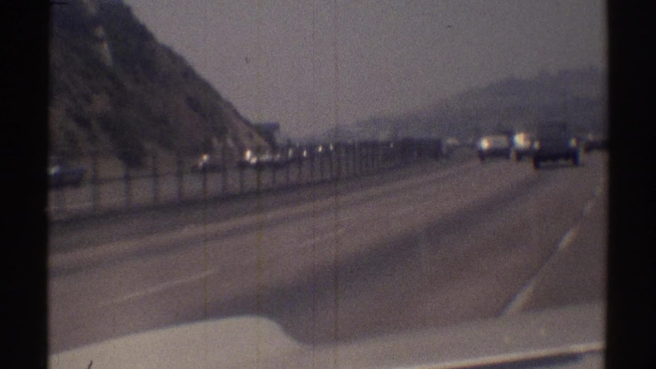 Forward view along a multi-lane highway beside a steep hillside and roadside fence.