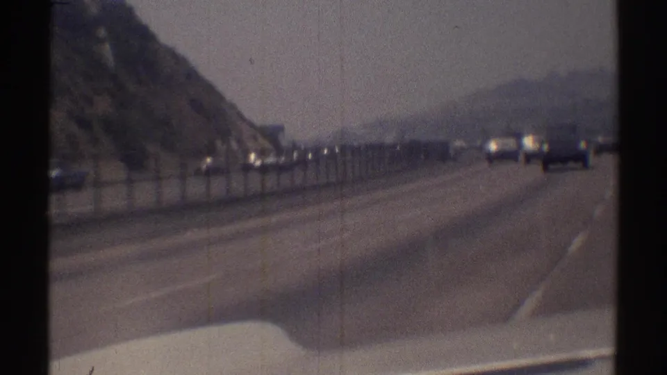 1969 Woodland Hills highway view from a moving vehicle, with lanes ahead, hillside on the left, fence, and distant traffic.