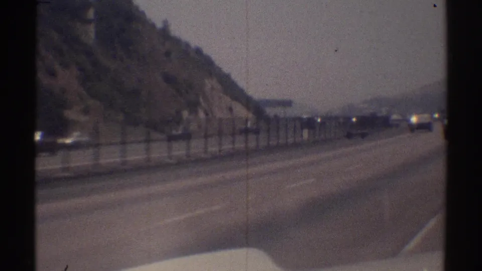 Vehicle-view footage on a Woodland Hills, California highway, showing a hillside edge, metal fence, and period traffic ahead.