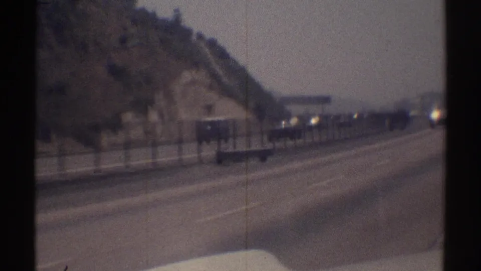 1969 highway travel view with a large vehicle near the left side, distant cars, and a steep hillside along the road.