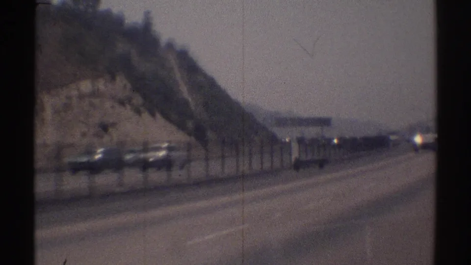 Woodland Hills highway scene from 1969, with classic cars, a rocky hillside, roadside fencing, and lanes stretching ahead.