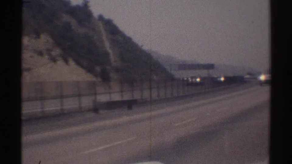 1969 Woodland Hills highway view from inside a moving vehicle, with hillside, fencing, and cars ahead in soft 8mm focus.