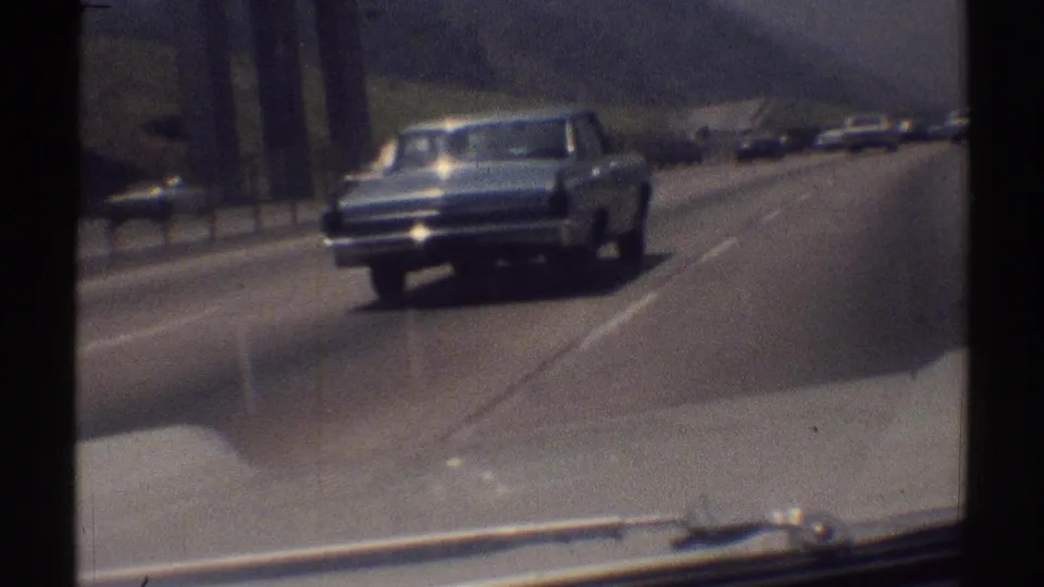 A light-colored classic sedan continues along the Woodland Hills highway, with roadside structure, hillside, and distant cars in view.