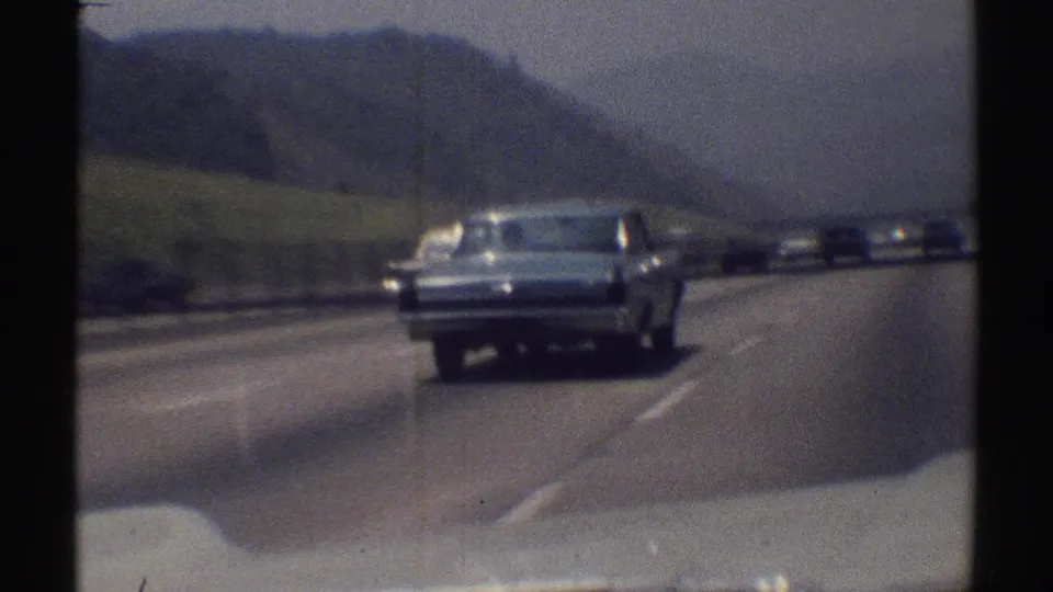 The blue sedan moves farther along the Woodland Hills highway, with several cars visible under a hazy 1969 sky.