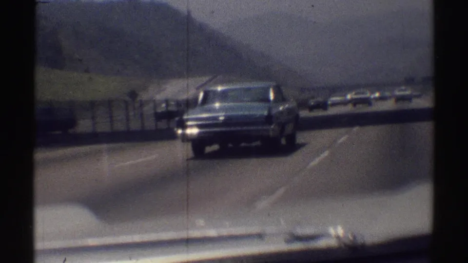 A close passing view of the blue sedan on a 1969 Woodland Hills highway, seen from inside another moving vehicle.