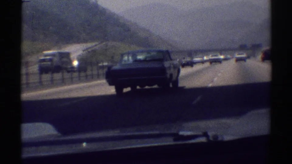 The blue sedan recedes on the Woodland Hills highway, framed by traffic, roadside hills, and the lower edge of the camera car windshield.