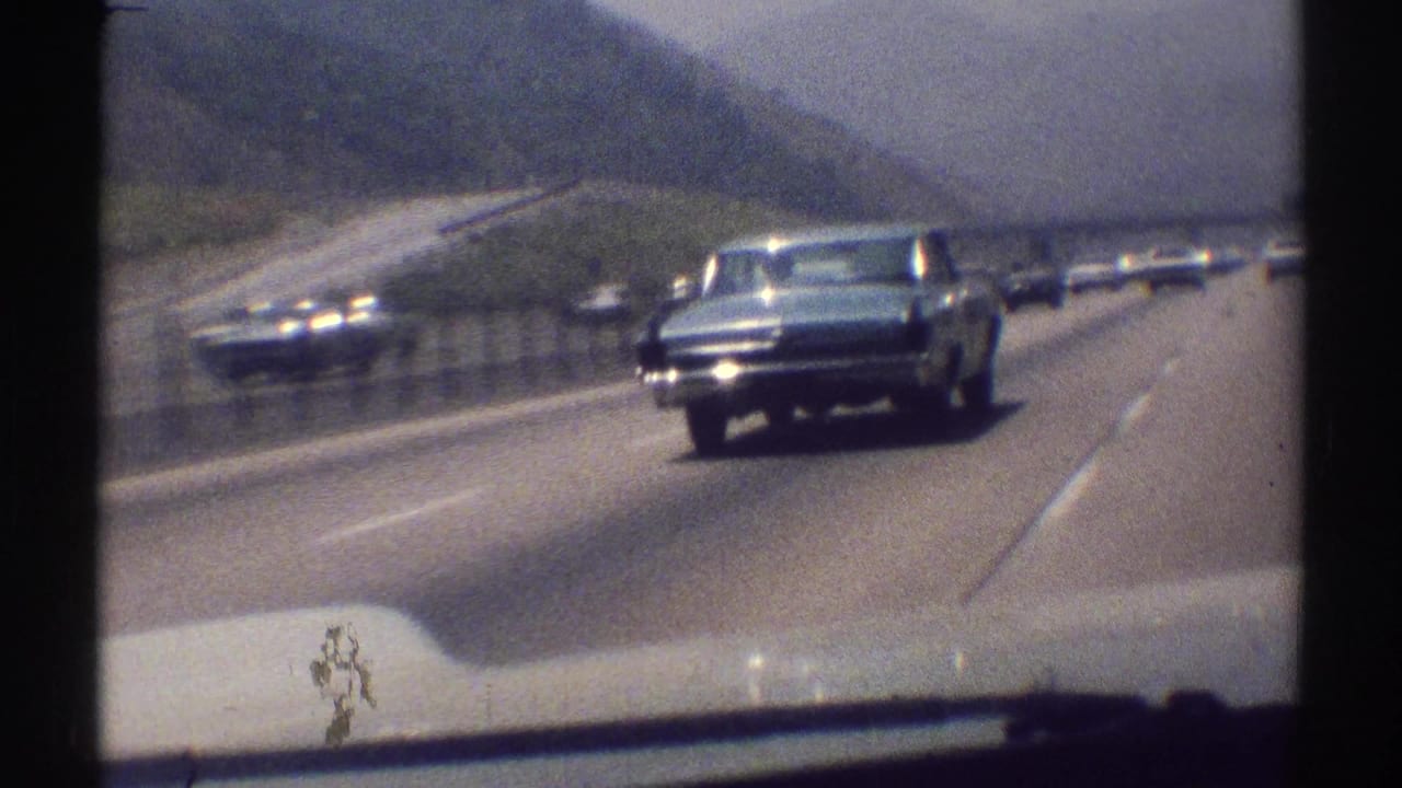 1969 Woodland Hills highway view from a moving car, with a dark sedan approaching amid traffic and hazy hills.