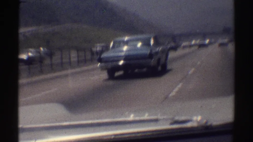 Dark 1960s sedan continues along a Woodland Hills highway, filmed through a soft 8mm passenger viewpoint.