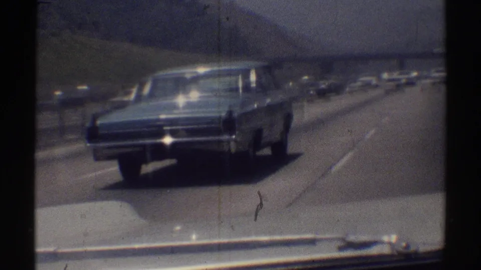 Close passenger-side view of a dark classic sedan on a Woodland Hills highway, with traffic and hills beyond.