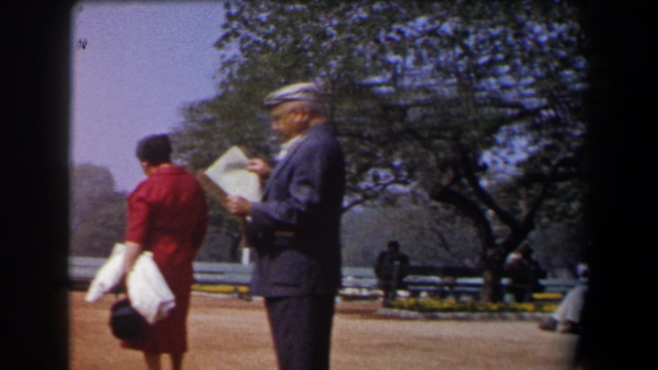 Woman in red dress and older man with papers stand beside benches under trees.
