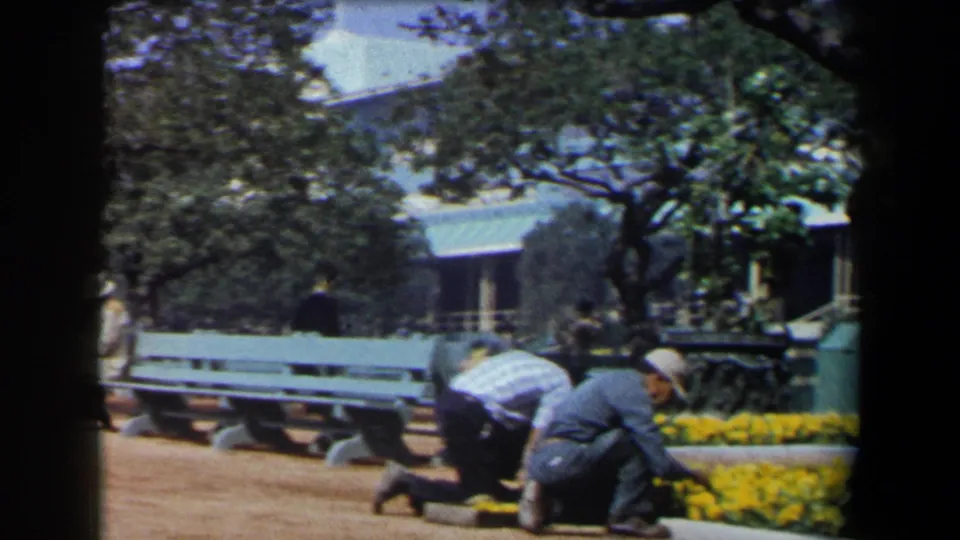 1958 California park frame showing a gardener bending near yellow flower beds, with benches, trees, and a small structure partly visible behind.