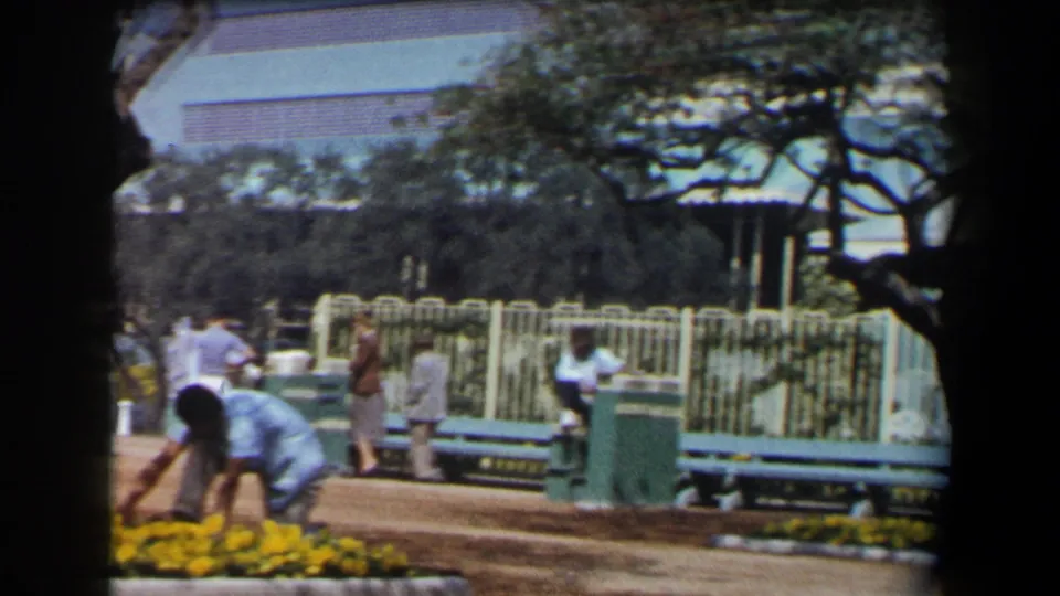 The frame shows a worker caring for yellow flowers in a California park, with benches, fencing, and strolling visitors behind.
