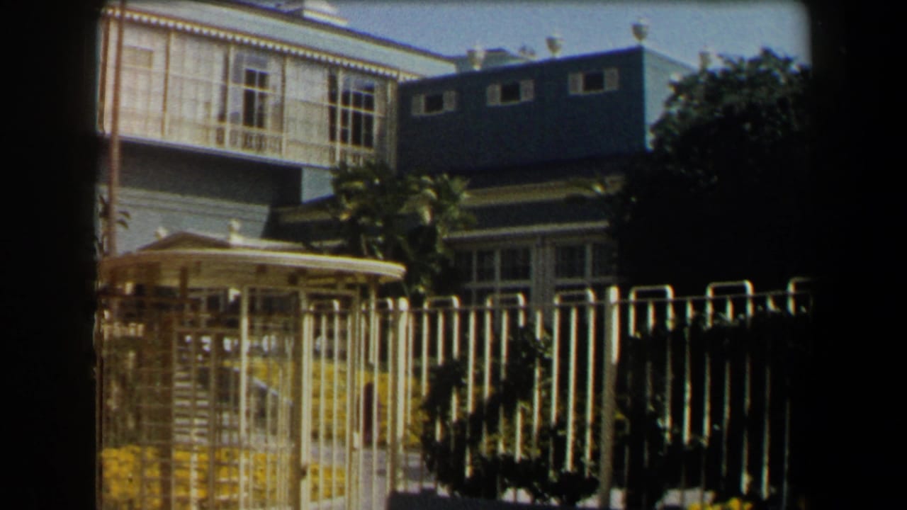 1958 California home-movie frame showing a blue two-story house with white trim, garden flowers, and a picket fence.