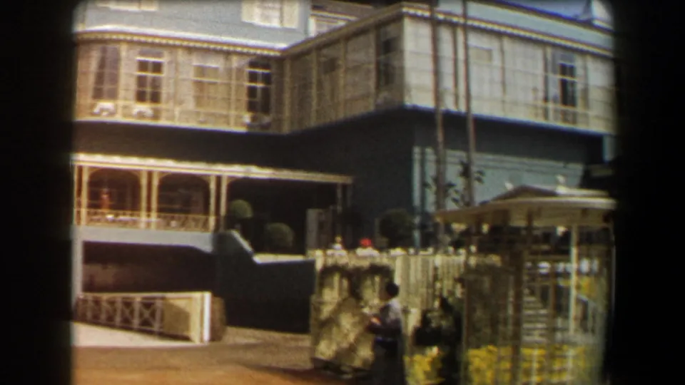 1958 California frame with the blue house porch, white fencing, yellow flowers, and people beside the garden gate.