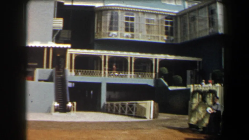 Wider 1958 California home-movie frame showing the blue house exterior, porch railings, blue benches, driveway, and people at the right edge.