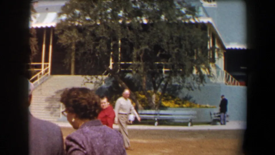 1958 California home-movie frame showing adults walking through a sunny exterior area with benches, stairs, a tree, and a pale blue wall.