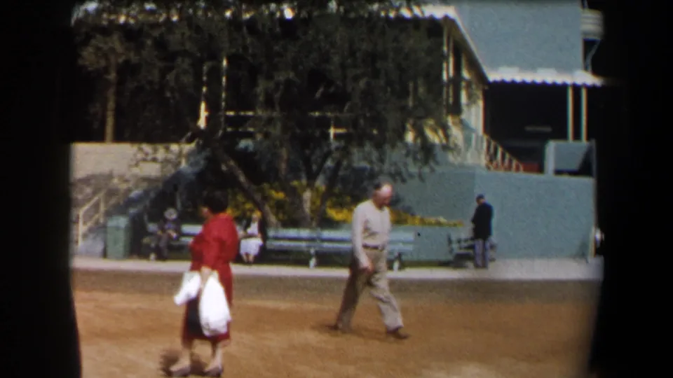 1958 California 8mm frame with pedestrians crossing a sunlit paved area near benches, a large tree, and a pale blue building exterior.