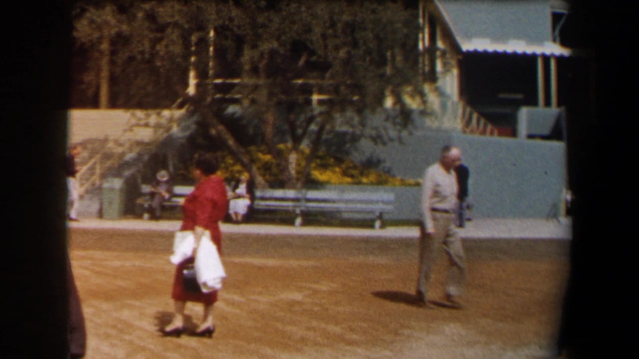 Woman in a red dress walks across a dirt path as a man in a light suit passes near benches.