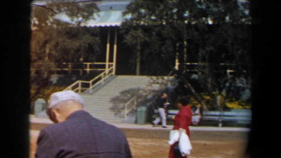 1958 California outdoor frame with a capped man in the foreground and pedestrians near benches, stairs, and a striped awning.