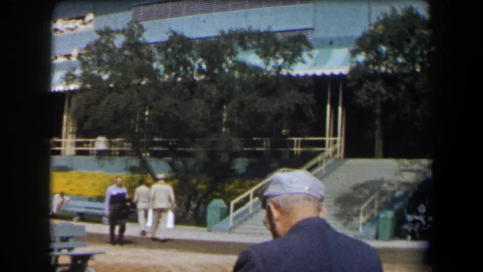 1958 California home-movie view of visitors near benches, yellow planting beds, stairs, and a shaded building entrance.