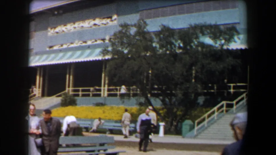 1958 California exterior frame showing pedestrians, benches, trees, railings, stairs, and a public building with striped awnings.