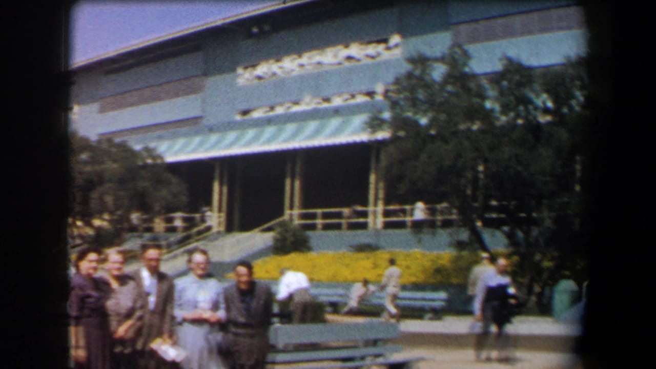 1958 California home-movie frame of adults posing near a large mid-century building with awnings, benches, flowers, and pedestrians.