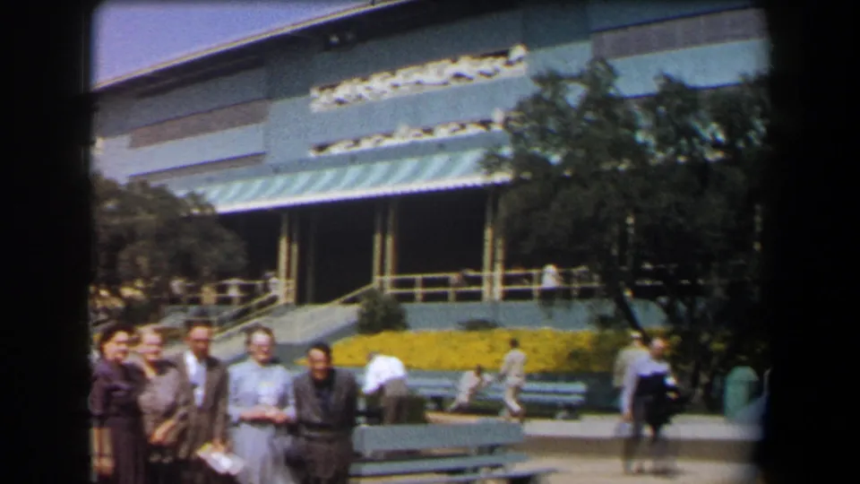 1958 California home-movie frame of adults posing near a large mid-century building with awnings, benches, flowers, and pedestrians.