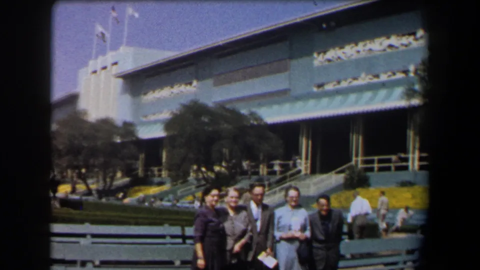 Five adults pose outside a large California building in 1958, with flags, striped awnings, green benches, and flower beds behind them.