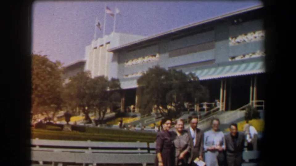 1958 California home-movie view of adults grouped near benches and yellow flowers outside a large building with flags and awnings.