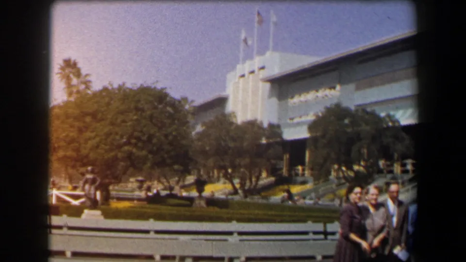 Adults are partly cropped at the edge of a 1958 California home-movie frame, with benches, trees, flags, and a large modern building visible.