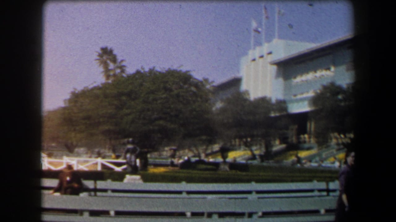 1958 California home-movie view of landscaped public grounds, a large building facade, benches, trees, and visitors in daylight.