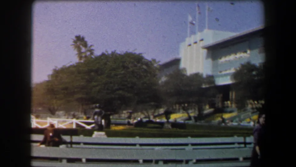1958 California home-movie view of landscaped public grounds, a large building facade, benches, trees, and visitors in daylight.
