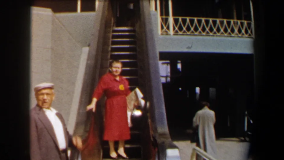 A woman in a red dress descends a mechanical stair at a California public venue while a cap-wearing man stands nearby.