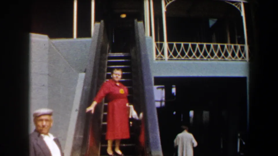 The woman in a red dress continues down the mechanical stair, holding the rail as a man in a cap stands in the foreground.