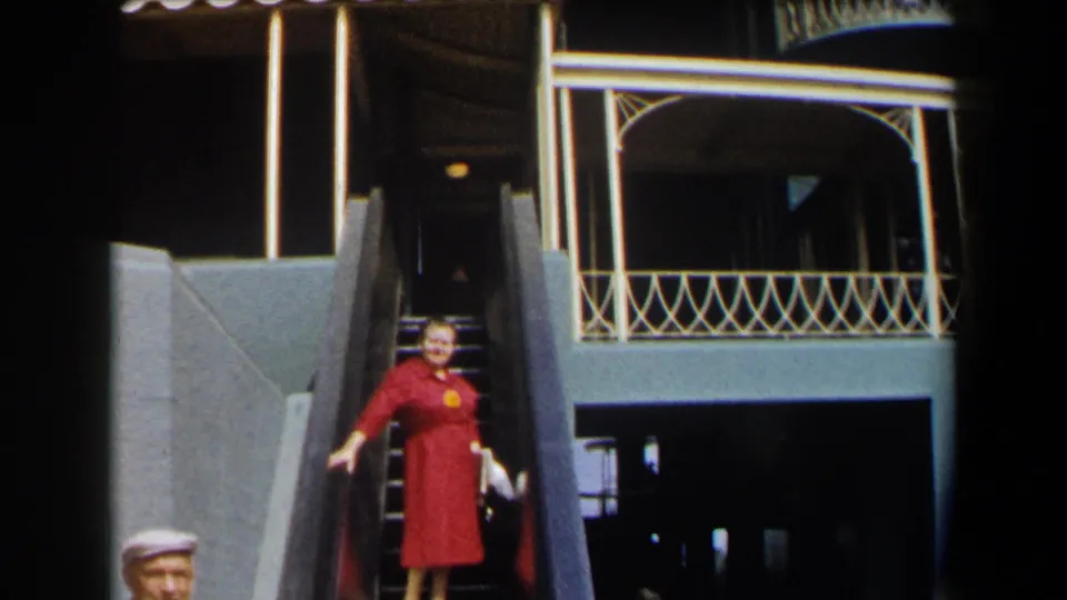 Near the end of the clip, the woman in red descends the mechanical stair in front of open railings and a shadowed building entry.