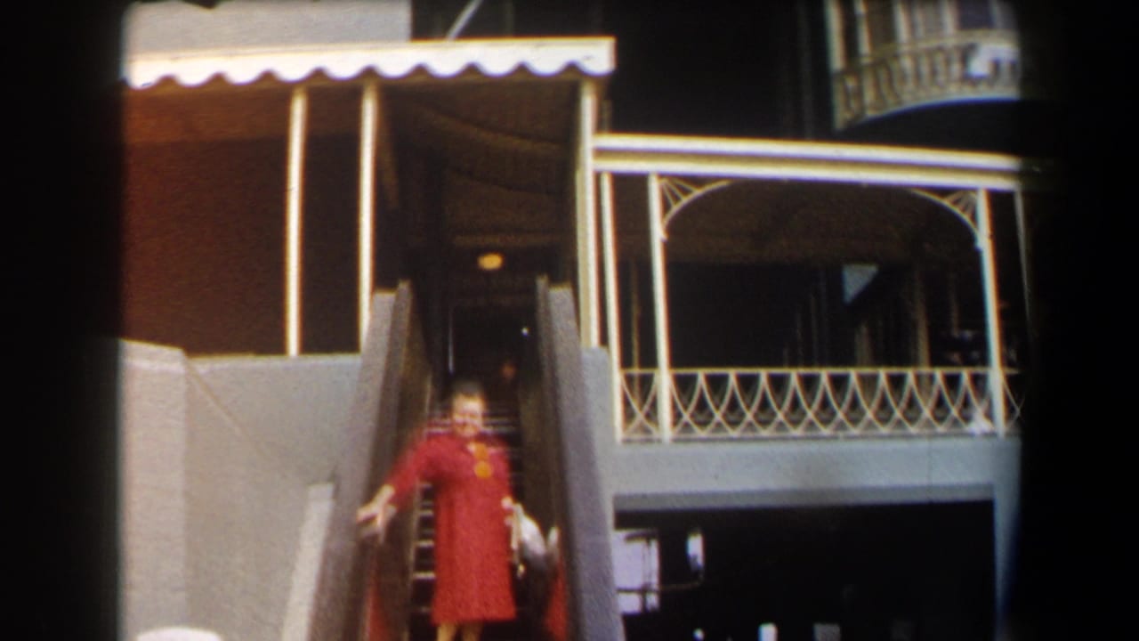 In 1958 California racetrack footage, a woman in a red dress waves from a stairway near a balcony and awning.