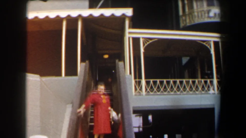 In 1958 California racetrack footage, a woman in a red dress waves from a stairway near a balcony and awning.