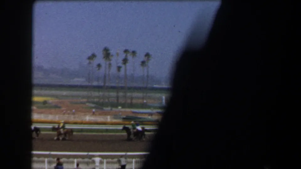 A handheld 8mm view of the racetrack shows horses in motion beyond railings, with palm trees and haze in the background.
