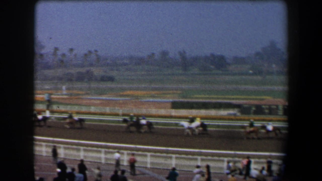 Racehorses with jockeys pass along a dirt track behind white railings as spectators watch.