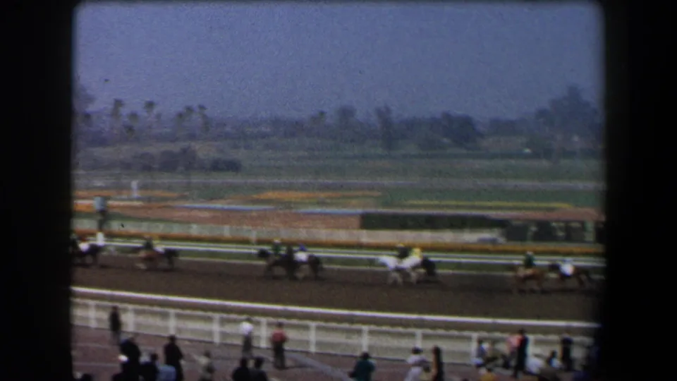 1958 California 8mm view of jockeys riding racehorses on a dirt track with spectators behind white railings.