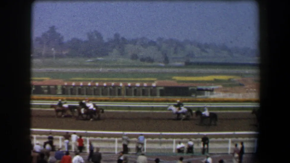 Soft 8mm frame of jockeys and horses rounding a dirt racetrack in California, with railings and spectators nearby.